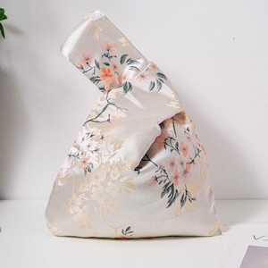 May include: A light beige fabric knot bag featuring a floral pattern in pink, peach, and gold. The bag has a distinctive knot closure. The floral design is a repeating pattern of blossoms and leaves. Suitable for carrying small items.