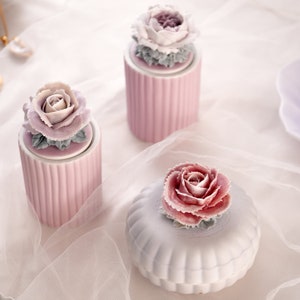 Op de afbeelding: Drie keramische containers met bloemmotieven. De containers zijn in roze en witte tinten. De containers staan op een wit oppervlak met een doorschijnende witte stofoverlay.