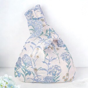 May include: A knotted fabric bag with a floral pattern in shades of blue and green on a cream background. The bag features a unique handle design and a soft, textured appearance, suitable for carrying items.