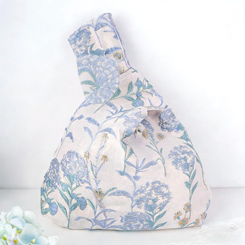 May include: A knotted fabric bag with a floral pattern in shades of blue and green on a cream background. The bag features a unique handle design and a soft, textured appearance, suitable for carrying items.