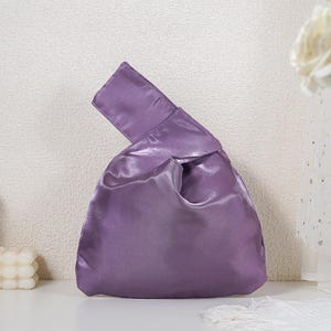 May include: A solid purple satin handbag featuring a Japanese-style knot handle design. The bag has a smooth, lustrous finish with a single asymmetrical handle loop that pulls through the other side to close the main compartment of the pouch.