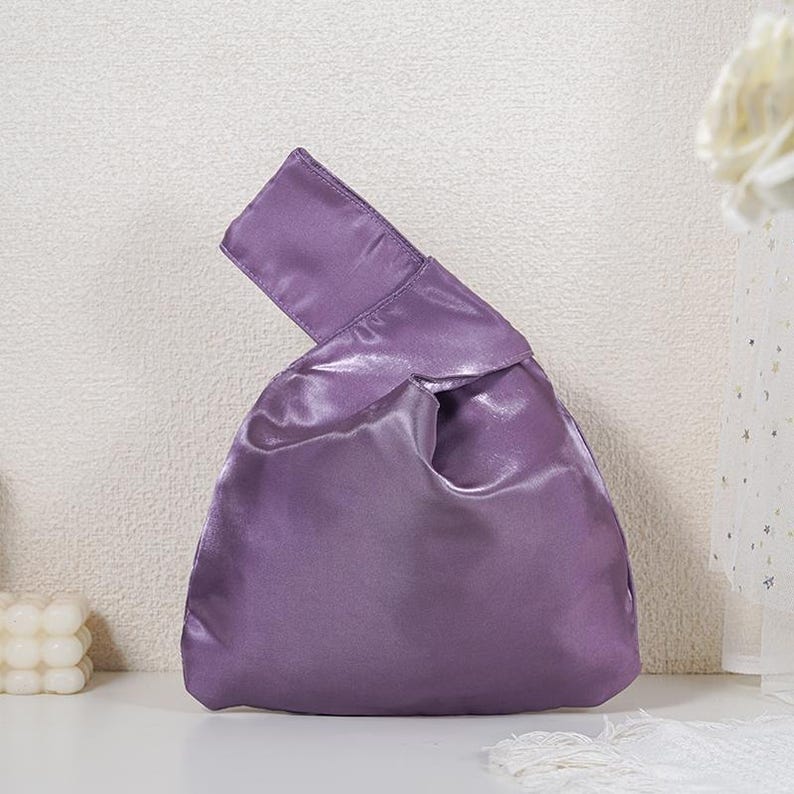 May include: A solid purple satin handbag featuring a Japanese-style knot handle design. The bag has a smooth, lustrous finish with a single asymmetrical handle loop that pulls through the other side to close the main compartment of the pouch.