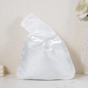 May include: A solid white satin fabric bag featuring a Japanese-style knot handle design. The bag has a smooth, reflective surface with a triangular shape and a single handle loop that is longer than the other, allowing it to fold over.