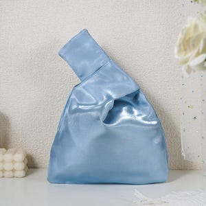 May include: A light blue satin handbag featuring a single integrated handle design where one side is longer than the other, creating an asymmetrical silhouette. The fabric has a smooth, reflective finish with soft folds and a structured base.
