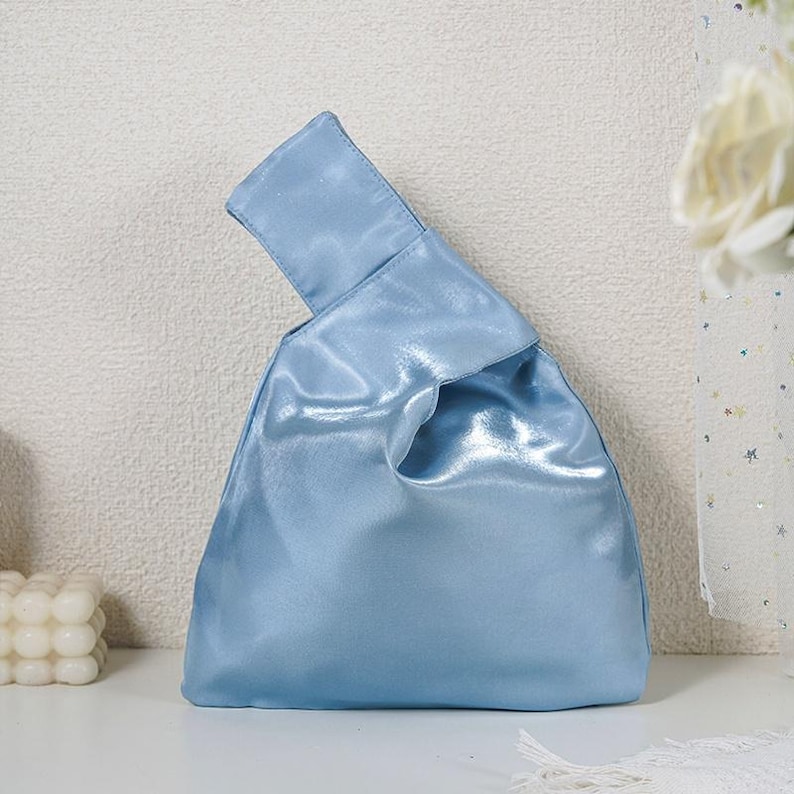 May include: A light blue satin handbag featuring a single integrated handle design where one side is longer than the other, creating an asymmetrical silhouette. The fabric has a smooth, reflective finish with soft folds and a structured base.
