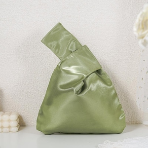 May include: A sage green satin handbag featuring a Japanese-style knot handle design. The bag has a smooth, lustrous finish and a triangular silhouette, with one handle longer than the other to allow for a secure, looped closure at the top.