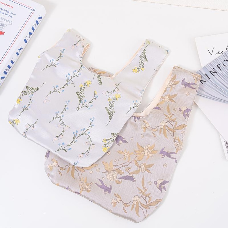 May include: Two satin tote bags with floral patterns. One bag is light grey with blue and yellow flowers. The other is tan with gold and purple floral and bird designs. Both bags have a knotted handle design.