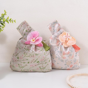May include: Two fabric pouches with floral patterns and orchid decorations. One has a pink orchid and green tassel, the other a peach orchid and orange tassel. The pouches have a light grey background with pink floral patterns.