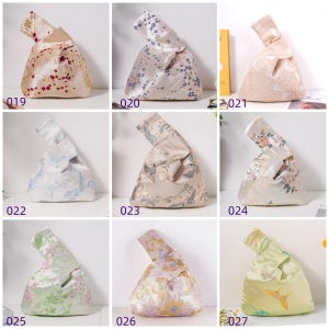 May include: A collection of nine Japanese knot bags in various colors and patterns. The bags are made of satin-like fabric and feature floral, bird, and abstract designs. Each bag has a unique color scheme, including cream, blue, pink, and gold.