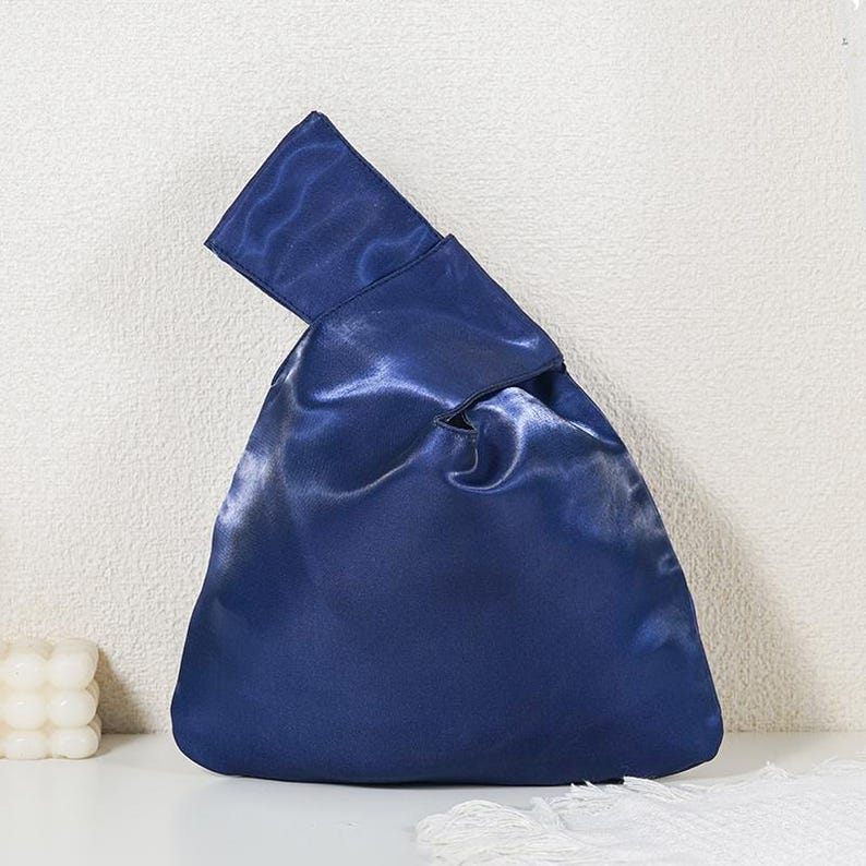 May include: A dark blue satin knot bag with a smooth, reflective finish. The bag features a triangular silhouette with one handle longer than the other, designed to loop through the shorter handle to close the opening at the top of the bag.