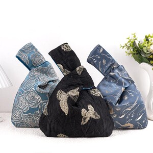 May include: Three decorative knot bags in blue and black. The blue bags feature floral and botanical patterns, while the black bag has a butterfly design. Each bag has a knotted handle and a contrasting blue interior.