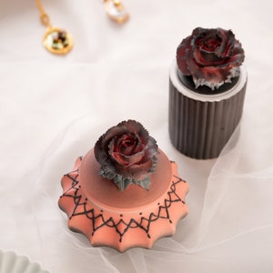 Può includere: Due scatole decorative con accenti di rose. La scatola a sinistra è rosa con un motivo di pizzo nero e una rosa rossa in cima. La scatola a destra è marrone con una rosa rossa in cima.