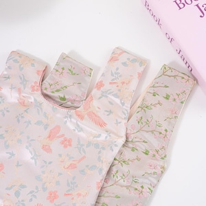 May include: Two reusable shopping bags with a floral and bird pattern. The bags are made of a silky, light pink fabric with a repeating design of pink birds, flowers, and leaves in shades of blue and cream.