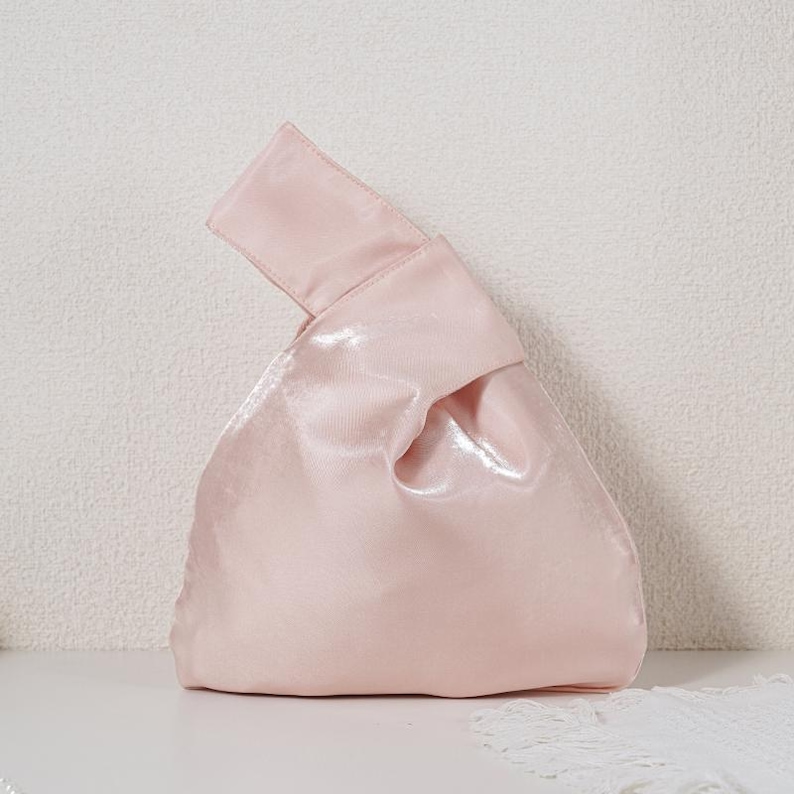 May include: A pale pink handbag with a soft, shimmering finish. The bag features a unique knot-style handle where one side is longer than the other, creating an asymmetrical silhouette. The material has a subtle sheen that reflects light across its surface.