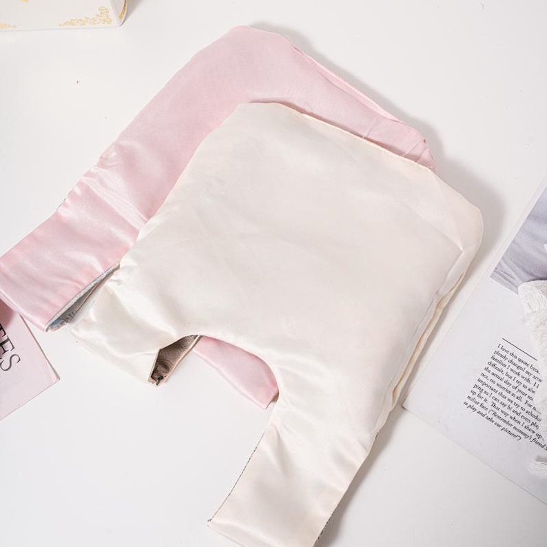 May include: Two satin sleep masks, one light pink and one cream, are displayed. The masks have a rectangular shape with a long, narrow strap extending from one side. The fabric has a slight sheen, suggesting a smooth texture.