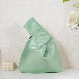 May include: A light mint green handbag with a smooth, satin-like finish. The bag features a unique asymmetrical silhouette with one handle longer than the other, allowing the longer handle to loop through the shorter one for a secure closure.