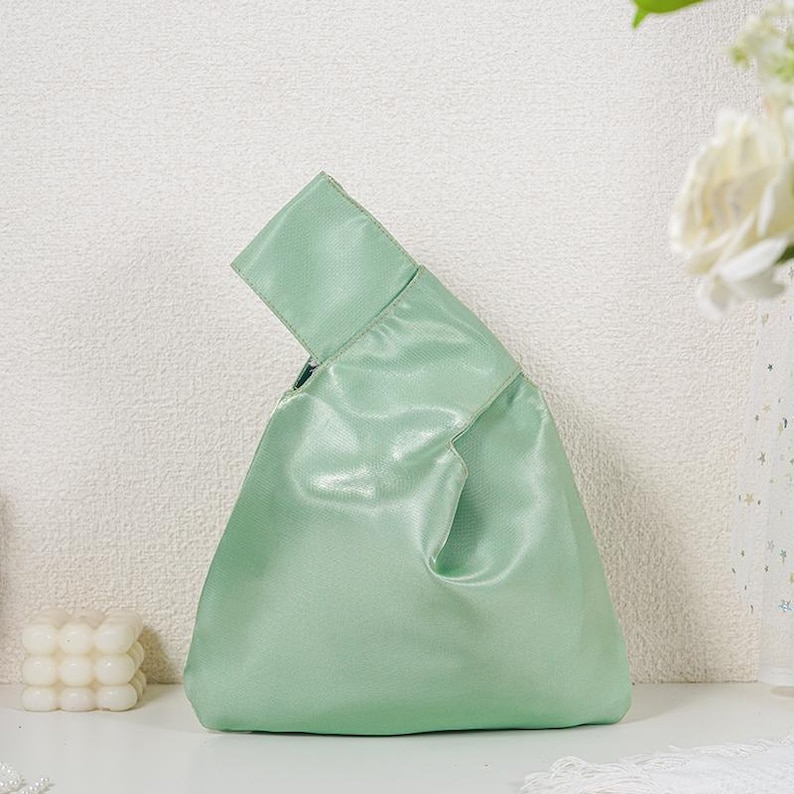 May include: A light mint green handbag with a smooth, satin-like finish. The bag features a unique asymmetrical silhouette with one handle longer than the other, allowing the longer handle to loop through the shorter one for a secure closure.