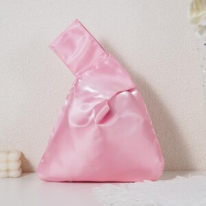 May include: A light pink satin handbag featuring a Japanese-style knot handle design. The bag has a triangular silhouette with a smooth, reflective fabric surface and a single integrated handle formed by one side being longer than the other.