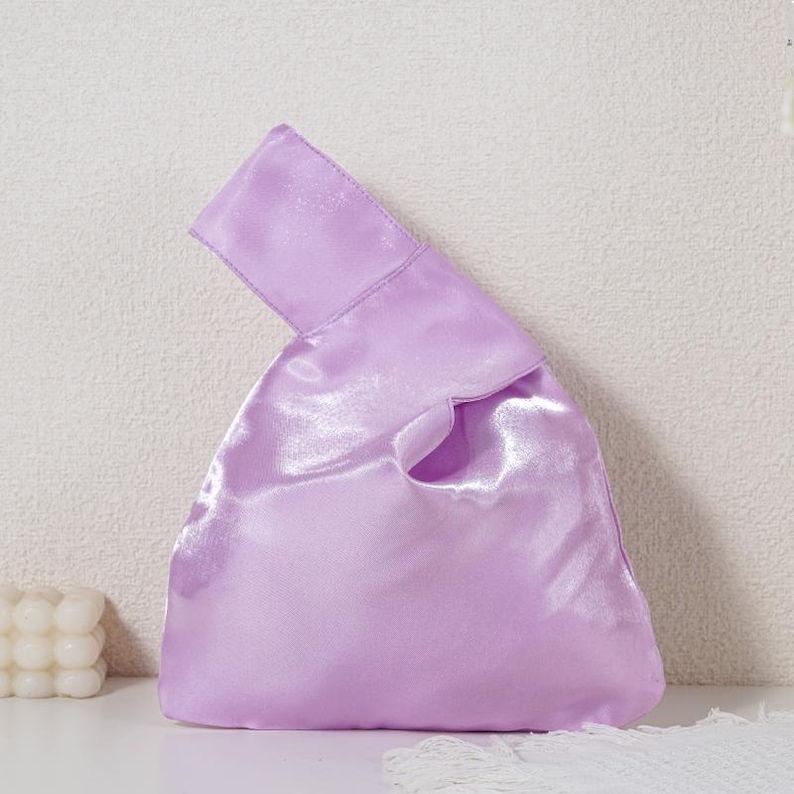 May include: A light purple satin knot bag with a smooth, reflective finish. The handbag features a single integrated handle design where one side is longer than the other, allowing the longer strap to pull through the shorter one to close.