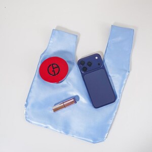 May include: A light blue satin knot-handle bag featuring a round red compact with a black GA logo, a dark blue smartphone with a triple-lens camera system, and a metallic lipstick tube resting on its surface against a plain white background.