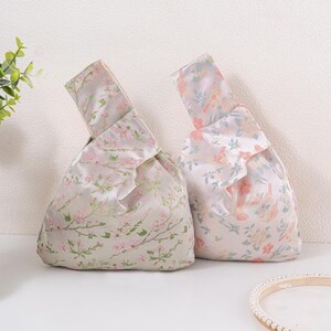 May include: Two floral-patterned bags with a knotted handle. The bags are made of a silky fabric with a light grey background and pink and green floral designs. The bags are a unique accessory.