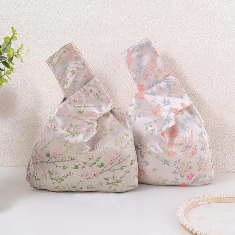 May include: Two floral-patterned bags with a knotted handle. The bags are made of a silky fabric with a light grey background and pink and green floral designs. The bags are a unique accessory.