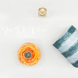 Op de afbeelding: Een blauwe en witte gestreepte keramische mok met de tekst "Joie De Vivre" in witte cursieve letters. Een klein doorzichtig glazen flesje met een gouden dop en een perzikkleurige en gele bloem zijn ook in de afbeelding.
