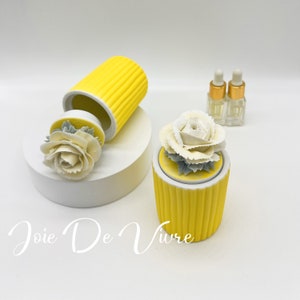 May include: A yellow ceramic diffuser with a white ceramic rose on top. The diffuser has a ribbed design and is sitting on a white surface. Two small glass bottles with gold caps are in the background. The text "Joie De Vivre" is written in cursive on the surface.