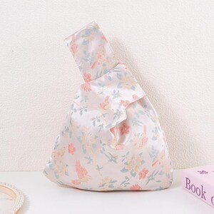 May include: A white satin handbag with a floral pattern in pastel colours. The design features birds and flowers in shades of pink, blue, and yellow. The bag has a knotted handle and a triangular shape.