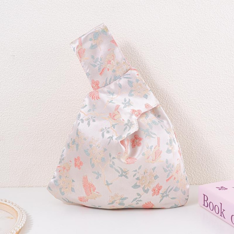 May include: A white satin handbag with a floral pattern in pastel colours. The design features birds and flowers in shades of pink, blue, and yellow. The bag has a knotted handle and a triangular shape.