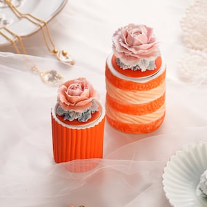 Op de afbeelding: Twee oranje keramische potten met bloemendeksels. De deksels zijn versierd met roze en witte bloemen. De potten hebben een getextureerde oranje afwerking.