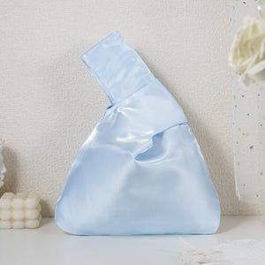 May include: A light blue satin handbag featuring a Japanese-style knot design with one handle longer than the other, allowing the longer handle to loop through the shorter one to close the bag. The fabric has a smooth, reflective finish.