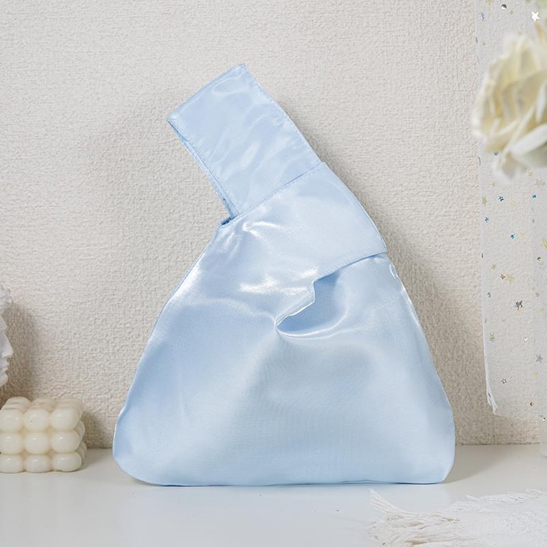 May include: A light blue satin handbag featuring a Japanese-style knot design with one handle longer than the other, allowing the longer handle to loop through the shorter one to close the bag. The fabric has a smooth, reflective finish.