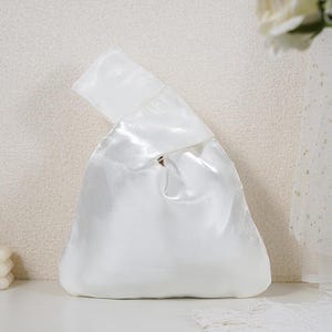 May include: A white satin knot bag featuring a smooth, reflective finish and a single integrated handle design. The handbag has a triangular silhouette with a gathered top opening, creating a soft, draped appearance with a subtle sheen.