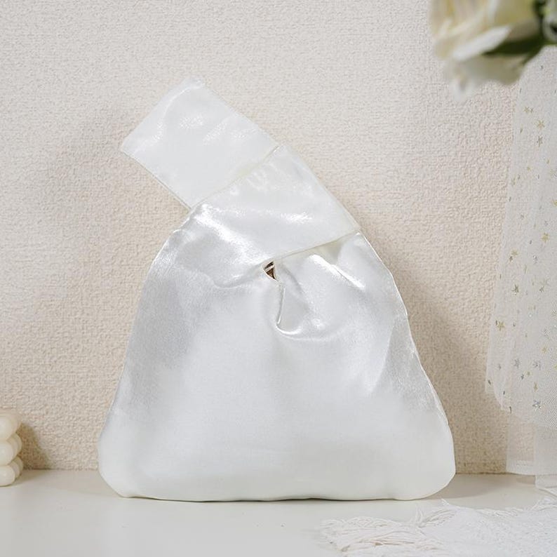 May include: A white satin knot bag featuring a smooth, reflective finish and a single integrated handle design. The handbag has a triangular silhouette with a gathered top opening, creating a soft, draped appearance with a subtle sheen.