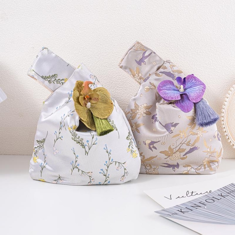 May include: Two fabric bags with floral patterns and orchid decorations. One is light grey with blue and green flowers, the other is light purple with gold flowers and birds. Each bag has a handle.