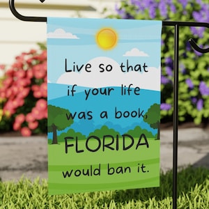 May include: A garden flag with a blue sky, white clouds, a yellow sun, and green trees. The text on the flag reads: "Live so that if your life was a book, FLORIDA would ban it."