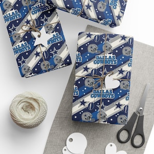 May include: Gift-wrapped presents with Dallas Cowboys themed wrapping paper. The paper features a repeating pattern of stars, football helmets, and the team name in blue, white, and gray. Each gift is tied with twine and a decorative tag.