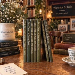 Op de afbeelding: Een verzameling groene boeken met harde kaft getiteld The Tale of Jemima Puddle-Duck, Peter Rabbit, Ginger and Pickles, Mrs. Tittlemouse, Benjamin Bunny, Two Bad Mice en Appley Dapply's Nursery Rhymes, op een houten tafel met een mok.