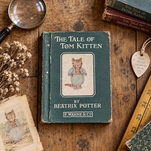 May include: A vintage green hardback book titled The Tale of Tom Kitten by Beatrix Potter, featuring a central illustration of a kitten in blue clothing. The book is surrounded by a magnifying glass, dried flowers, and a wooden ruler.