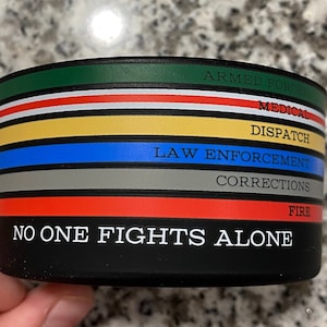 Peut inclure: Une tasse noire et blanche avec un motif de rayures multicolores. Les rayures représentent différentes professions de premiers intervenants : forces armées, médical, répartition, application de la loi, corrections et pompiers. Le texte « NO ONE FIGHTS ALONE » est imprimé sur la tasse.