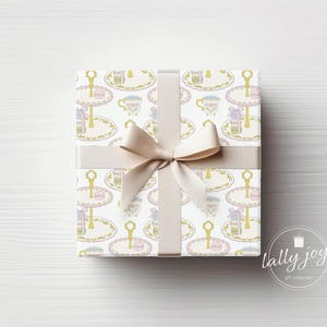 May include: A white gift box with a beige ribbon tied in a bow. The box is wrapped in paper with a repeating pattern of teacups and tiered cake stands. The text "lally joy!" is printed on the bottom right corner of the image.