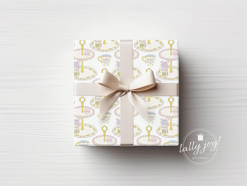 May include: A white gift box with a beige ribbon tied in a bow. The box is wrapped in paper with a repeating pattern of teacups and tiered cake stands. The text "lally joy!" is printed on the bottom right corner of the image.