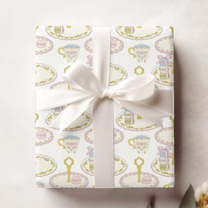 May include: White gift wrap with a repeating pattern of teacups, saucers, and macarons in pastel colors. The gift is tied with a white satin ribbon.