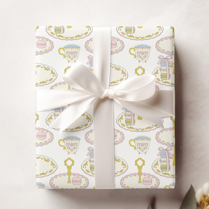 May include: White gift wrap with a repeating pattern of teacups, saucers, and macarons in pastel colors. The gift is tied with a white satin ribbon.