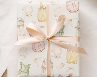 May include: A white gift wrap with a pink ribbon. The wrapping paper features a repeating pattern of watercolor illustrations of baby clothes, including dresses, bonnets, and bloomers.