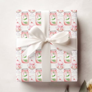May include: White gift wrap with a repeating pattern of pink and green watercolor illustrations of jars and cherries. A white satin ribbon is tied around the gift box.