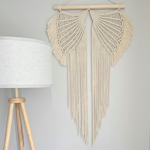 Op de afbeelding: Crèmekleurige macramé wandhanger met een waaiervormig ontwerp, opgehangen aan een houten deuvel. De wandhanger heeft ingewikkelde knopen en franjes. Een houten statieflamp met een witte lampenkap staat op de voorgrond.