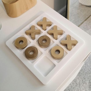 Puede incluir: Un juego de tres en raya de madera con cruces y círculos de color latón sobre una cuadrícula blanca. Una caja rectangular de madera con una ranura está en el fondo. El juego está sobre una superficie blanca.