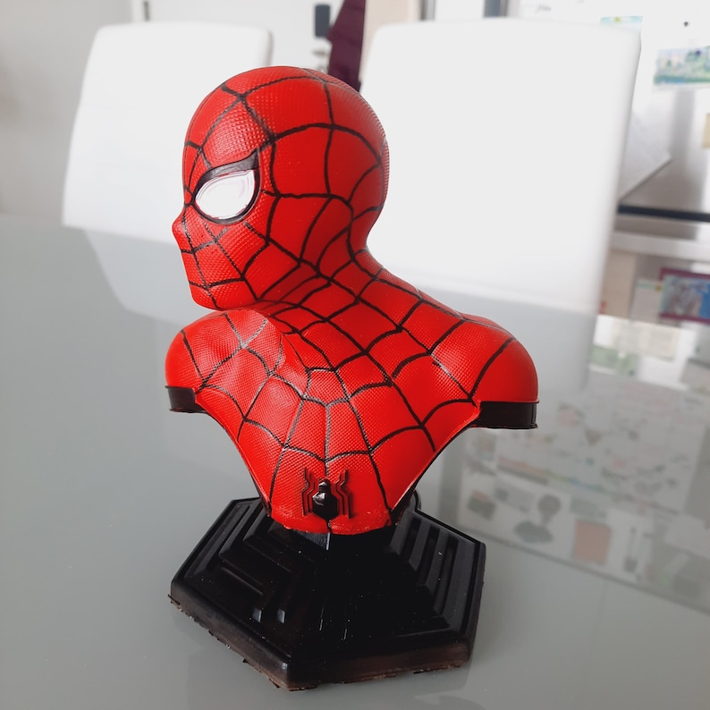 May include: A red and black Spider-Man bust figurine. The bust features the character's mask with white eye lenses and a web pattern. The figurine is mounted on a black, geometric base.